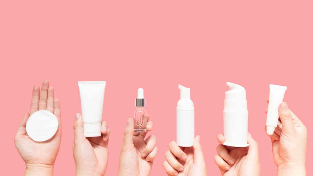 Woman hands holding facial cotton pad, foam, essential oil, serum, lotion and eye cream packaging on pink background. 