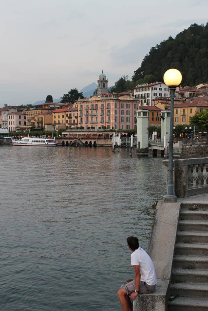 A man sits on a concrete wall looking towards the shoreline of Bellagio Italy on Lake Como