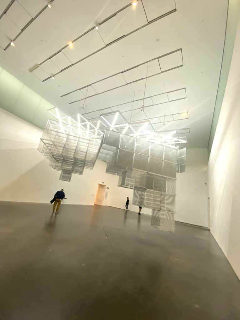 A giant sculpture made of squares hangs from the ceiling of a large exhibit room
