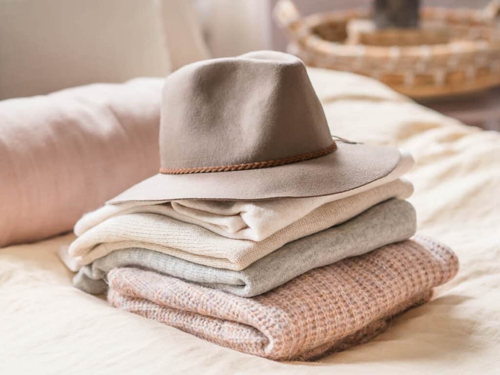 a brown hat on top of a pile of folded neutral colored sweaters 