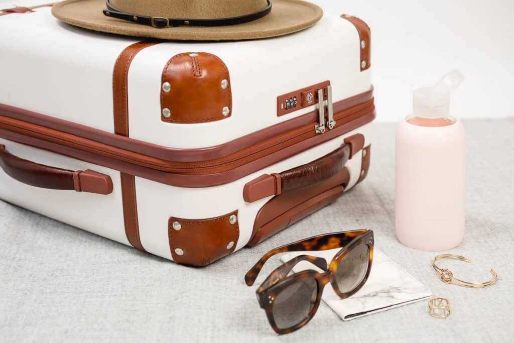 sunglasses and a pink travel toiletry bottle placed outside of a suitcase with a hat on top of it