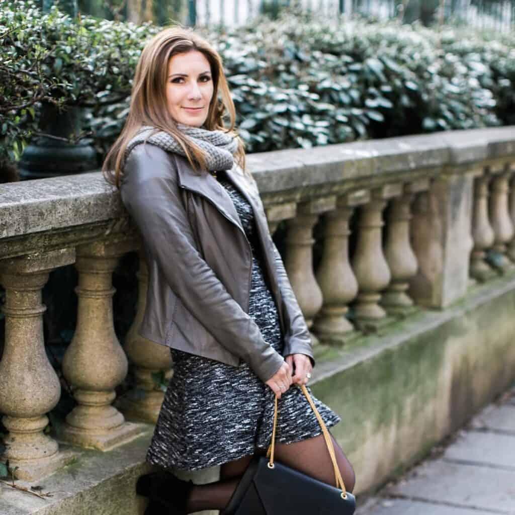 Jessica leaning against a wall wearing a scarf and holding a purse.